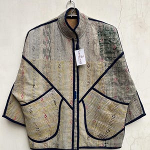 May include: A quilted jacket featuring a patchwork design in muted tones of beige, yellow, and green with dark blue trim. The jacket has a high collar, wide sleeves, and two large front pockets. A white tag labeled Lareeba Gifts is attached.