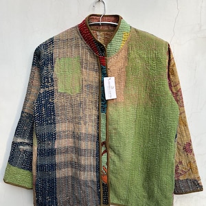 Peut inclure: Une veste à manches longues composée de pièces de tissu matelassé multicolores dans des tons de vert, bleu et marron. La veste présente un col officier, un liseré marron sur les bords et une étiquette rectangulaire blanche marquée LARESSA GIFTS.