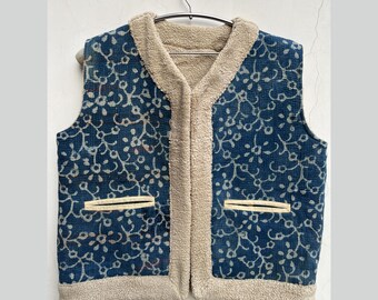 May include: A blue and white patterned vest with a beige fleece lining. The vest has a zipper closure and two pockets.