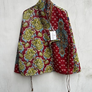 May include: A quilted wrap jacket featuring a vibrant red base with green and blue floral patterns on one side and a geometric print on the other. The garment includes thin brown tie closures and a white tag labeled LARISSA GIFTS.