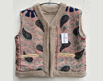 May include: A sleeveless vest with a pink paisley pattern and a beige sherpa lining. The vest has two front pockets with yellow trim and a tag that reads "US LABELS GIFTS". The vest is hanging on a metal hanger.
