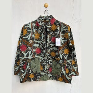 May include: A dark-colored jacket with an intricate floral pattern in red, yellow, and green. The jacket has a collared neckline, long sleeves, and two front pockets. A tag is attached to the jacket. The jacket is hanging on a wall.
