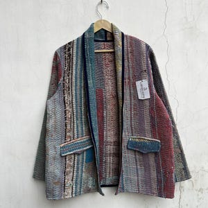 Puede incluir: Una chaqueta de manga larga con un patrón de retazos multicolor con rayas verticales en tonos rojo, azul, gris y bronceado. La chaqueta tiene un cuello chal azul oscuro, dos bolsillos delanteros con solapas y una etiqueta blanca que dice LARISSA GIFTS.