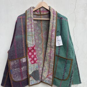 Puede incluir: Una chaqueta de manga larga abierta por delante con un diseño de patchwork de texturas y colores variados, incluyendo morado, verde y rosa. La prenda tiene dos grandes bolsillos delanteros con ribete marrón y una etiqueta blanca LARESSA GIFTS.