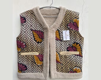 May include: A reversible vest with a geometric pattern and leaf-like designs in shades of burgundy, gold, and brown. The vest has a beige, fuzzy lining and trim, with two front pockets. A tag reads "LARESSA GIFTS".
