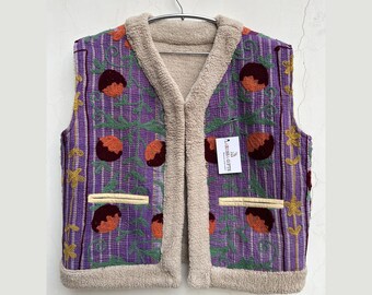 May include: A sleeveless purple vest with a floral pattern and a beige sherpa lining. The vest features a vertical striped design with embroidered orange and brown fruit motifs. The edges and collar are trimmed with beige sherpa. A tag is attached to the vest.
