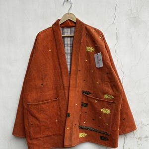 May include: A burnt orange quilted jacket featuring a shawl collar, long sleeves, and two front patch pockets. The fabric has a textured surface with small yellow and black embroidered motifs. A white tag is attached to the front chest area.