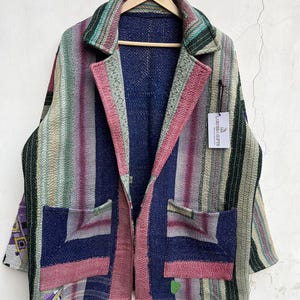 May include: A long-sleeve jacket featuring a patchwork design with vertical stripes in shades of pink, green, blue, and cream. The garment has a notched collar, two large front pockets, and a white tag labeled LARESSA GIFTS hanging from the lapel.
