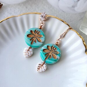 Könnte beinhalten: Ein Paar türkisfarbene Ohrringe mit Blattdesign in Gold. Die Ohrringe sind aus Polymer Clay gefertigt und haben einen weißen Blatt-Anhänger, der an jedem Ohrring hängt.
