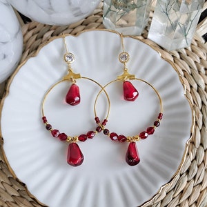 Könnte beinhalten: Ein Paar goldene Creolen mit herabhängenden roten tropfenförmigen Kristallen. Die Creolen sind mit kleinen roten Perlen verziert.
