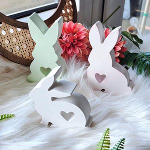 May include: Three wooden bunny figurines with a heart cutout on their chest. The bunnies are painted in pastel colours: green, pink, and white. The bunnies are sitting on a white fluffy surface.