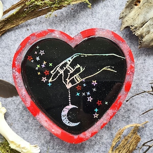May include: Heart-shaped resin tray with a black interior featuring a hand holding a crescent moon and stars design. The tray has a red border with glitter accents, ideal for holding small items.
