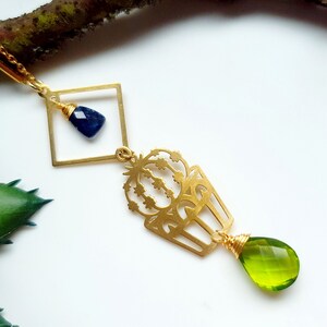 Könnte beinhalten: Eine goldfarbene Halskette mit Anhänger, die einen quadratischen Rahmen mit einem blauen Edelstein, einen kaktusförmigen Anhänger und einen grünen, tropfenförmigen Edelstein aufweist. Die Halskette befindet sich auf weißem Hintergrund mit einem Ast und einer grünen Pflanze.