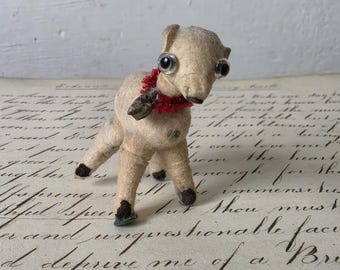 Puede incluir: Figura de cordero de estilo vintage con ojos grandes y un collar rojo. El cordero es beige con pezuñas negras y una oreja pequeña y marrón. La figura está sobre una superficie con escritura manuscrita.