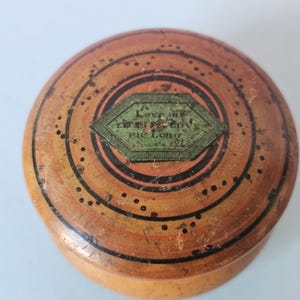 Può includere: Una scatola rotonda in legno con coperchio. La scatola è dipinta con cerchi concentrici nei toni dell'arancione e del marrone, con dettagli neri. Un'etichetta verde e bianca al centro recita "Love me the Longest".