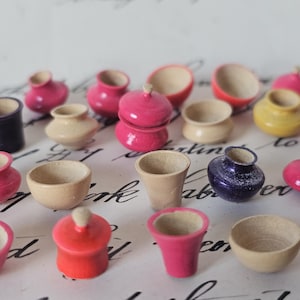 Puede incluir: Una colección de miniaturas de ollas y cuencos de madera pintados a mano en varias formas y colores. El surtido incluye tonos rosa, rojo, amarillo y madera natural. Algunos tienen tapas, otros son cuencos o tazas abiertos.