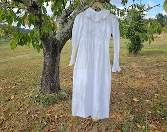 Puede incluir: Un vestido blanco de algodón con cuello de encaje y mangas largas. El vestido tiene una cintura fruncida y está plisado en todo su largo. Está colgado de una rama de árbol en una zona de hierba con hojas caídas.