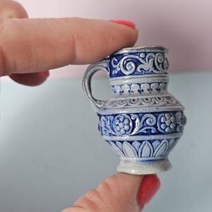 May include: A miniature blue and white ceramic pitcher with a handle. The pitcher features intricate floral and scrollwork designs. The color scheme is a combination of light gray and deep blue, with a slightly textured surface.