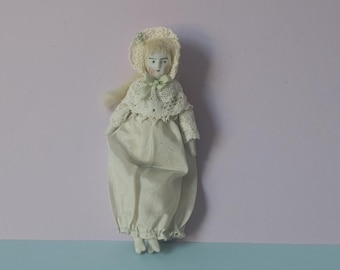 May include: A porcelain doll wearing a white dress and a white crocheted bonnet with green accents. The doll has blonde hair and blue eyes.