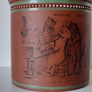 Puede incluir: Una jarra de terracota con una banda verde y blanca alrededor de la parte superior. La jarra presenta una ilustración en blanco y negro de una fiesta musical con gatos tocando instrumentos. El texto "MUSICAL PARTY" está impreso en la jarra.