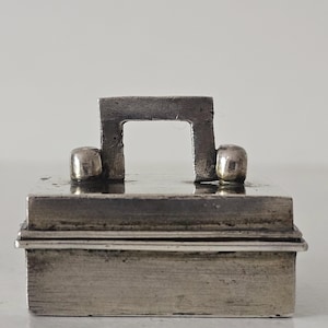 May include: A small, rectangular silver box with a handle and two rounded knobs. The handle is a square U-shape. The box has a hinged lid and appears to be made of metal. The box is likely used for storage.