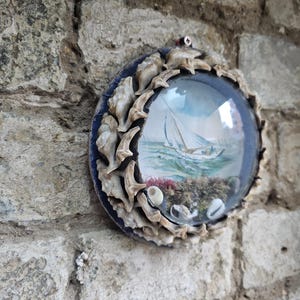 Puede incluir: Una decoración mural redonda de temática náutica. La obra de arte presenta un velero sobre un mar azul, encerrado en una cúpula de cristal. El marco está adornado con conchas marinas y se encuentra sobre una pared de piedra desgastada.