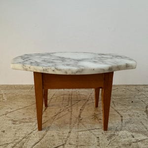 以下が含まれることがあります： 木製の脚と白い大理石の天板を持つ小さなテーブル。テーブルは独特の不規則な形をしており、薄い茶色の床の上に置かれています