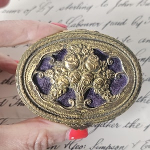 Op de afbeelding: Een antiek, ovaalvormig, goudkleurig metalen compact. De deksel heeft een reliëf bloemenontwerp met een centrale paarse fluwelen inzet. De compact is in een hand gehouden, met een handgeschreven document op de achtergrond.