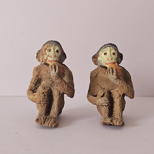 Puede incluir: Dos pequeñas figuras de monos vintage. Los monos son marrones con caras claras y sostienen un trozo de comida. Las figuras están sentadas con las piernas cruzadas.