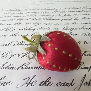 May include: A strawberry-shaped pincushion crafted from red fabric with embroidered green seed details. The top features a textured, light green fabric calyx with a small, protruding stem. The object is rounded and soft in appearance.