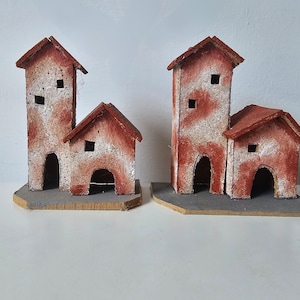Puede incluir: Dos juegos de casas en miniatura hechas de cartón pintado de marrón y blanco. Cada juego tiene dos casas, una más alta que la otra. Las casas tienen pequeñas ventanas y puertas.