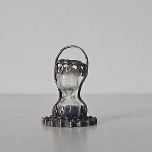 May include: A small, ornate hourglass with a clear glass bulb and a metal frame. The frame is decorated with intricate designs and has a dark bronze finish.