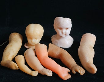 Puede incluir: Una colección de piezas de muñeca vintage, que incluyen una cabeza, brazos y piernas, hechas de plástico blanco roto. Las piezas están dispuestas sobre una superficie negra.