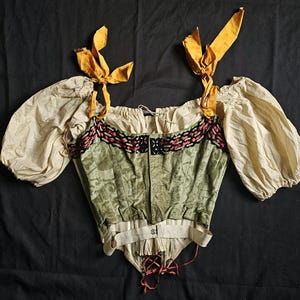 Op de afbeelding: Een lijfje in antieke stijl met crèmekleurige pofmouwen en een groen en crèmekleurig lijfje. Het lijfje heeft rode en zwarte afwerking en gele strikken op de schouders. Het kledingstuk ligt plat op een zwart oppervlak.