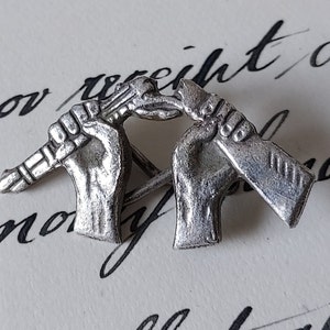 Op de afbeelding: Een paar zilverkleurige reverssieraden, elk met een hand die een wapen vasthoudt. De ene hand houdt een geweer vast, de andere een tomahawk.