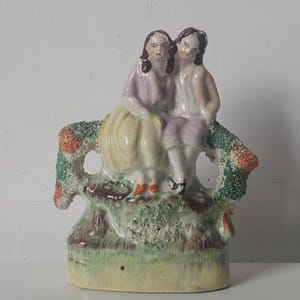 以下が含まれることがあります： 木の上に座っている2人の人物を描いた、ヴィンテージの磁器製置物。人物はパステルカラーの服を着ており、ベースは緑と黄色の色合いです。木の幹にはカラフルなテクスチャの詳細があります。