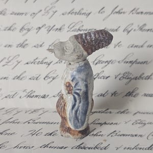 May include: A small, antique figurine of a person, possibly a soldier or a historical figure. The figurine is painted with muted colors, including brown, blue, and white. It has a detailed hat and clothing, suggesting a vintage or collectible item.