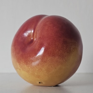 May include: A ceramic peach ornament with a glossy finish. The peach has a gradient of colours, transitioning from a deep red at the top to a yellow-orange at the bottom. The peach is a decorative item.