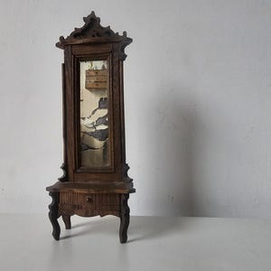 May include: A miniature antique wooden cabinet with a mirror. The cabinet is dark brown with ornate carvings and a small drawer. The mirror has a distressed appearance. The cabinet stands on four curved legs.