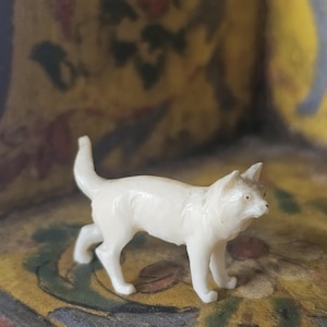 Könnte beinhalten: Eine kleine, weiße, geschnitzte Elfenbeinfigur eines Katers. Die Katze steht auf allen vieren und hat den Kopf nach rechts gedreht. Die Figur ist detailliert mit realistischen Merkmalen, einschließlich eines langen Schwanzes und spitzer Ohren.