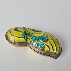 Könnte beinhalten: Eine kleine, gelbe, erdnussförmige Schmuckschatulle mit grünem Blumendesign. Die Schachtel ist aus Metall und hat eine glänzende Oberfläche.