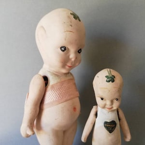 May include: Two vintage porcelain dolls with painted green shamrock accents. The larger doll has a pink fabric bodice and the smaller doll has a white fabric bodice with a heart-shaped label that reads "FUN-SUP."
