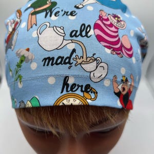May include: A blue scrub hat with white polka dots and a whimsical Alice in Wonderland theme. It features characters like the Mad Hatter, the Cheshire Cat, and the Queen of Hearts. The text "We're all mad here" is printed on the hat.