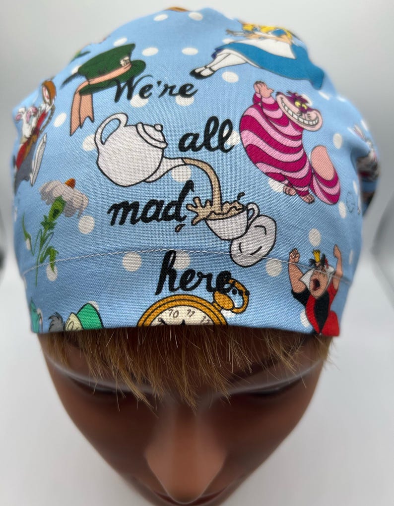 May include: A blue scrub hat with white polka dots and a whimsical Alice in Wonderland theme. It features characters like the Mad Hatter, the Cheshire Cat, and the Queen of Hearts. The text "We're all mad here" is printed on the hat.
