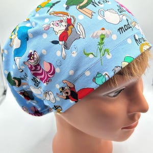 May include: A blue scrub hat with a white polka dot pattern and a whimsical Alice in Wonderland print featuring characters like the Mad Hatter, the White Rabbit, and the Cheshire Cat. The hat has the text "We're all mad here" printed on it.