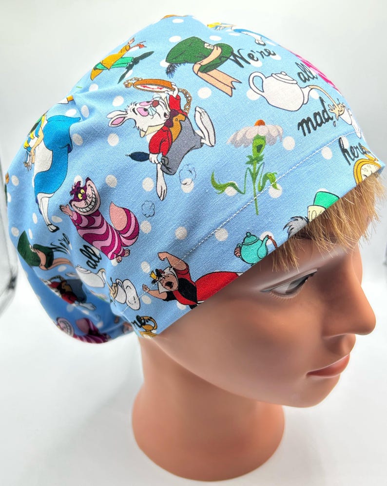 May include: A blue scrub hat with a white polka dot pattern and a whimsical Alice in Wonderland print featuring characters like the Mad Hatter, the White Rabbit, and the Cheshire Cat. The hat has the text "We're all mad here" printed on it.