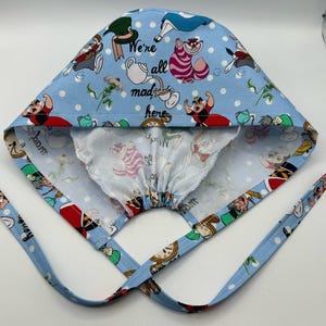 May include: A blue and white surgical cap with a print of characters from Alice in Wonderland. The cap has a drawstring and ties. The text "We're all mad here" is printed on the cap.