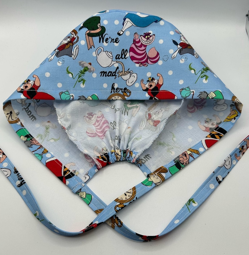 May include: A blue and white surgical cap with a print of characters from Alice in Wonderland. The cap has a drawstring and ties. The text "We're all mad here" is printed on the cap.