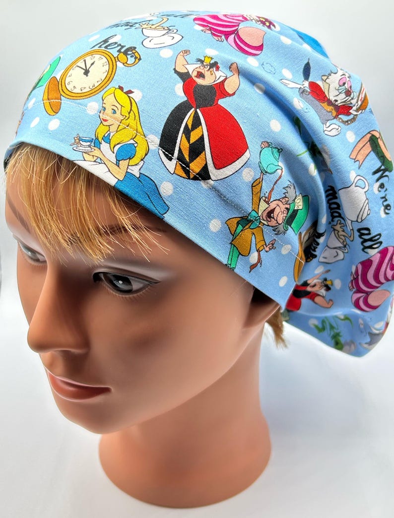May include: Blue scrub hat with a white polka dot pattern and a colorful Alice in Wonderland print featuring characters like the Queen of Hearts, Mad Hatter, and Alice. The text "We're all mad here" is printed on the hat.