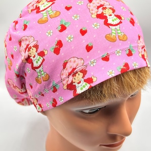 May include: Pink scrub cap featuring a repeating pattern of a cartoon character with red hair and a strawberry hat, strawberries, and white flowers. The cap is made of a soft fabric and is designed to cover the head.